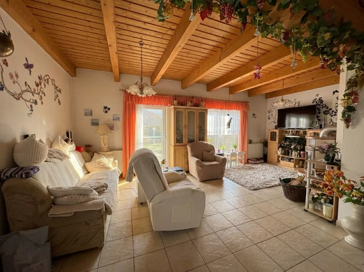 Wohnzimmer mit Sofas, Sesseln, gefliestem Boden, Holzbalkendecke und großen Fenstern zum Garten.