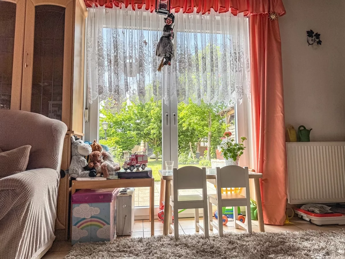 Spielecke im Wohnzimmer mit kleinem Kindertisch, zwei Stühlen und Spielzeug vor dem großen Fenster.
